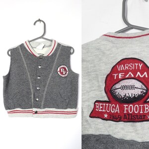May include: Gray sleeveless sweater vest with red and white trim. The vest has a button closure and a patch on the front that says "Varsity Team Beluga Football 1969 Allstar".