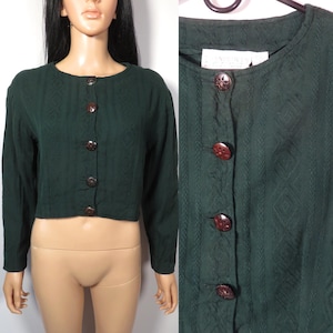 May include: A dark green cropped cardigan with a textured pattern and brown buttons. The cardigan has long sleeves and a round neckline.