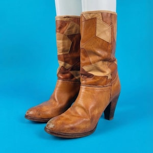 May include: Mid-calf boots featuring a patchwork design of various tan and brown leather segments. The boots have a rounded toe, a stacked block heel, and visible stitching along the seams of the leather panels throughout the shaft and foot.