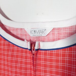 May include: Close-up of a red and white plaid jumpsuit with a white collar and blue trim. The zipper is red. The tag reads "KOMAR MADE IN U.S.A."