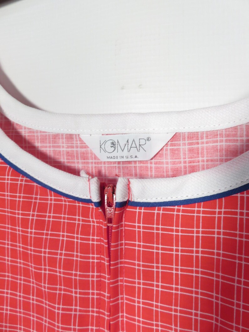May include: Close-up of a red and white plaid jumpsuit with a white collar and blue trim. The zipper is red. The tag reads "KOMAR MADE IN U.S.A."