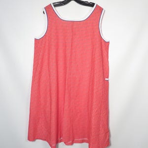 May include: A sleeveless, red gingham dress with white trim and a navy blue accent. The dress is hanging on a black hanger against a white background. The dress has a simple, classic design.
