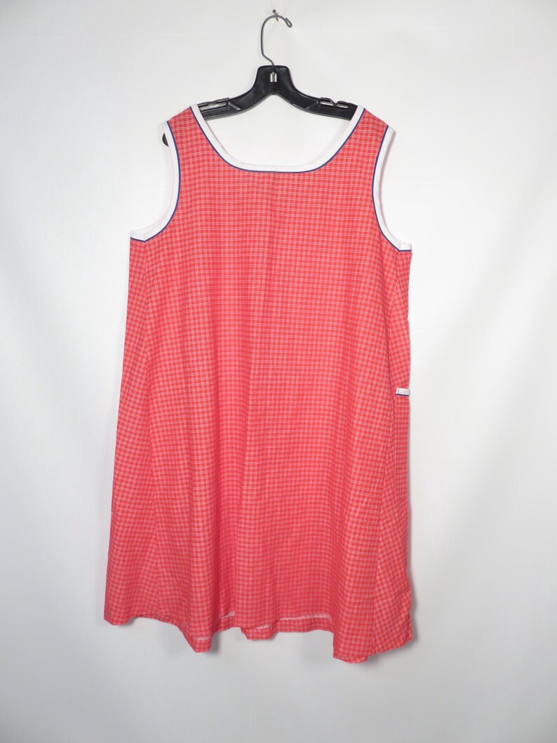 May include: A sleeveless, red gingham dress with white trim and a navy blue accent. The dress is hanging on a black hanger against a white background. The dress has a simple, classic design.