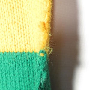 May include: Close-up of a knitted fabric with a yellow and green color scheme. The fabric is textured and has a loose weave. The edge of the fabric is frayed.