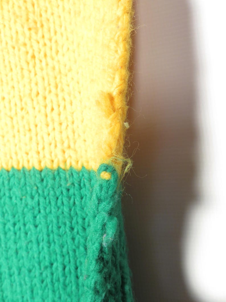 May include: Close-up of a knitted fabric with a yellow and green color scheme. The fabric is textured and has a loose weave. The edge of the fabric is frayed.