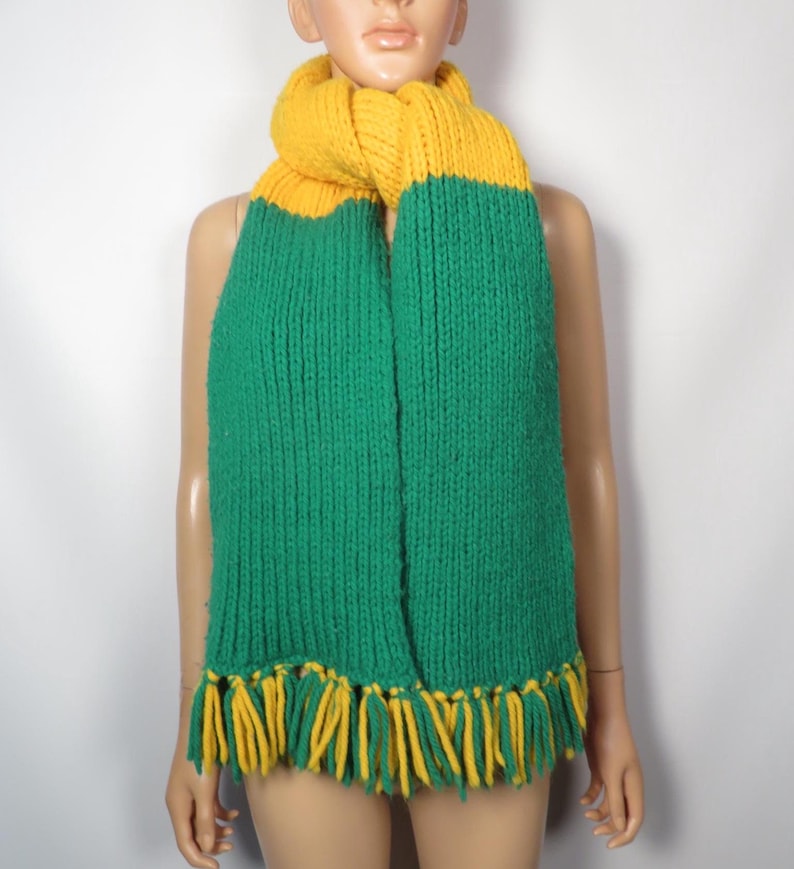 May include: A green and yellow knitted scarf with fringe. The scarf is made of a soft, chunky yarn and has a simple, classic design.