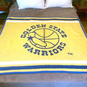 May include: A yellow and blue fleece blanket featuring the Golden State Warriors logo. The logo depicts a basketball with a star and the words "Golden State Warriors" surrounding it.