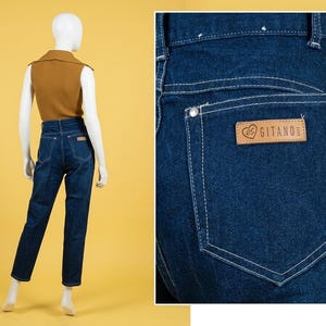 May include: Dark blue denim jeans featuring a high-waisted fit, white contrast stitching, and a tan leather patch on the back pocket with the text "es GITANO". The jeans are shown on a mannequin wearing a mustard yellow sleeveless ribbed top.