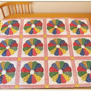 May include: A quilt featuring a grid of twelve circular flower motifs. Each flower consists of multi-colored fabric petals in red, blue, green, and yellow patterns on a white background. The grid is separated by red and white checkered sashing.