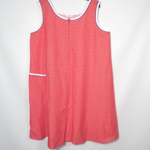 May include: A sleeveless, knee-length dress in a red and white checkered pattern. The dress features white trim with a navy blue border along the neckline and armholes. A small pocket with white trim is visible on the front. The dress is hanging on a black hanger.