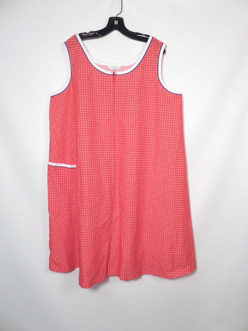 May include: A sleeveless, knee-length dress in a red and white checkered pattern. The dress features white trim with a navy blue border along the neckline and armholes. A small pocket with white trim is visible on the front. The dress is hanging on a black hanger.