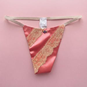 Könnte beinhalten: Ein korallenroter Satin-String mit diagonalen Einsätzen aus beiger Spitzenborte. Das Kleidungsstück verfügt über einen dünnen, elastischen beigen Bund und eine kleine, dekorative Blumenapplikation in der vorderen Mitte unter einem weißen Etikett.
