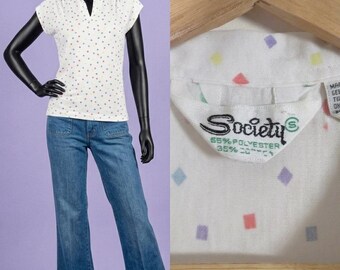 May include: A white short-sleeve polo shirt with a colorful diamond pattern and blue denim jeans. The shirt features a collar, a V-neckline, and a label reading Society, 55% polyester, 35% cotton. The jeans have two front pockets and a flared leg cut.