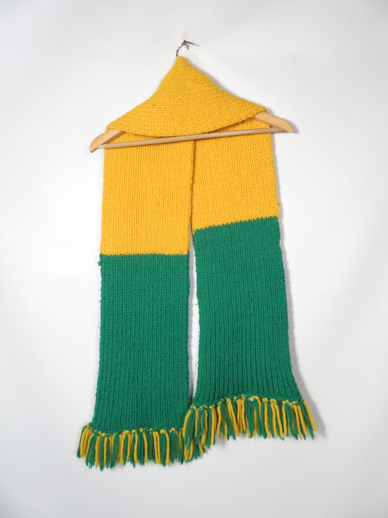 May include: A yellow and green knitted scarf with fringe on the ends. The scarf is hanging on a wooden hanger.