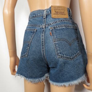 以下が含まれることがあります： フリンジヘムのブルーデニムショートパンツ。ハイウエストで、後ろにLevi'sのブランドパッチが付いています。