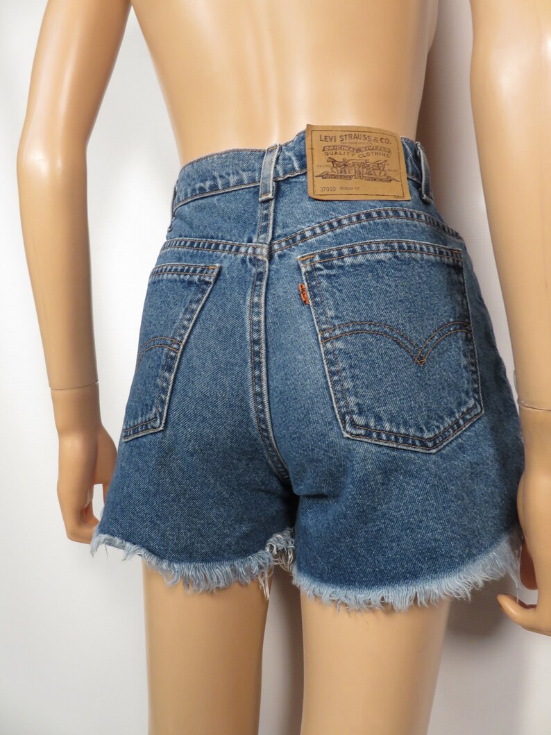 以下が含まれることがあります： フリンジヘムのブルーデニムショートパンツ。ハイウエストで、後ろにLevi'sのブランドパッチが付いています。