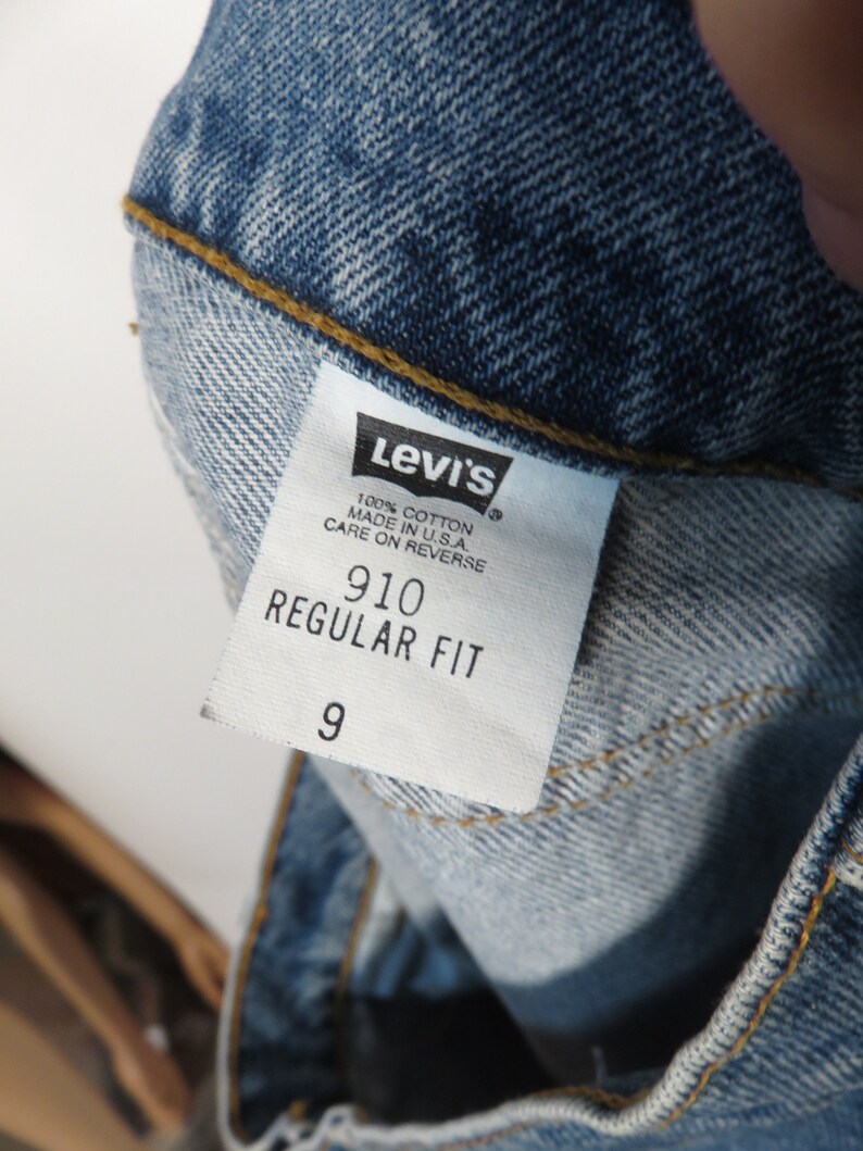 以下が含まれることがあります： 「Levi's」という文字、 「100% コットン」、「米国製」、「裏返してのお手入れ」、「910 レギュラーフィット」、「9」という文字が書かれた白い Levi's タグ。
