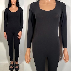 May include: A black jumpsuit with a scoop neckline and long sleeves. The one-piece garment is form-fitting and made of a stretchy material. The model is wearing black platform sandals.
