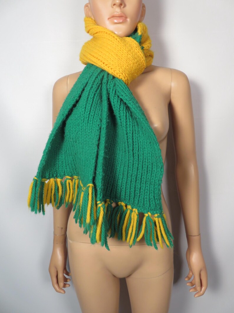 May include: A green and yellow knitted scarf with fringe. The scarf is made of a soft, chunky yarn and has a simple, classic design.