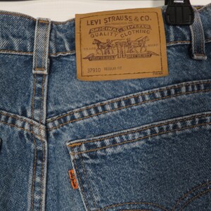 以下が含まれることがあります： Levi Strauss & Co. のブラウンレザーパッチが付いたブルーデニムのジーンズ。パッチには「Original riveted quality clothing」と「37910 regular fit」の文字が書かれています。