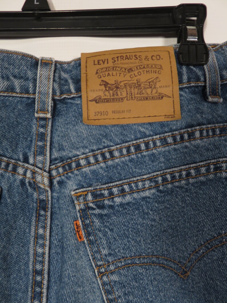 以下が含まれることがあります： Levi Strauss & Co. のブラウンレザーパッチが付いたブルーデニムのジーンズ。パッチには「Original riveted quality clothing」と「37910 regular fit」の文字が書かれています。