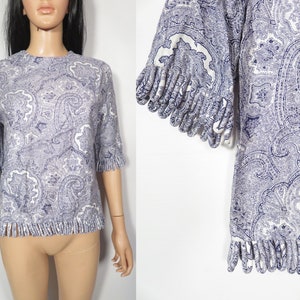May include: A white and blue paisley print top with a fringe trim around the neckline and sleeves. The top is made of a soft, lightweight fabric.