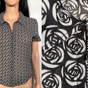 May include: Short-sleeved button-up shirt with a black and white rose pattern. The shirt has a collared neckline and a zipper closure. The fabric appears to be lightweight and semi-sheer.