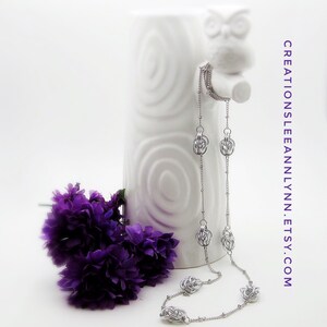 May include: A silver chain necklace with multiple silver knot-shaped charms. The necklace is displayed on a white ceramic owl figurine.