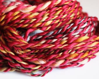 May include: A close-up of a skein of yarn with a mix of red, orange, yellow, and grey colours. The yarn is twisted and piled together in a circular shape.