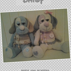 Op de afbeelding: Twee gehaakte knuffels in de vorm van honden, een blauw en beige en de andere roze en beige, met het woord "DAISY" erboven en de tekst "Sapie van Schepen" eronder.