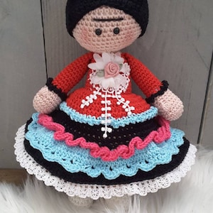 May include: A crocheted doll with black hair, a red dress, and a blue, pink, and white flower crown. The doll is wearing a black and white skirt with a blue and pink ruffle.