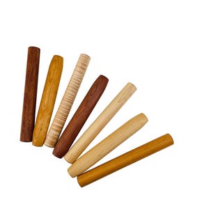 May include: A collection of seven cylindrical wooden dowels or pen blanks arranged in a fan shape. Each piece displays a unique wood grain pattern and color, ranging from light cream and golden yellow to medium brown and deep reddish-brown tones.