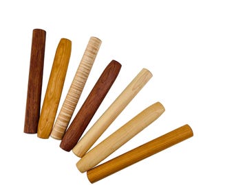 May include: A collection of seven cylindrical wooden dowels or pen blanks arranged in a fan shape. Each piece displays a unique wood grain pattern and color, ranging from light cream and golden yellow to medium brown and deep reddish-brown tones.