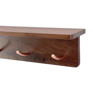 May include: A wall-mounted shelf made of dark brown wood with a visible grain pattern. The shelf features a flat top surface and a vertical back panel equipped with three curved, metallic copper-colored hooks for hanging items.
