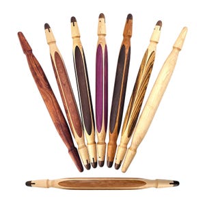 May include: A collection of seven handcrafted wooden rolling pins arranged in a fan shape with one additional rolling pin positioned horizontally below. Each tool features a tapered design with contrasting wood grains and colors including light, dark, and purple tones.