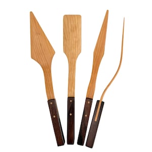 May include: A set of four kitchen utensils featuring light-colored wooden heads and dark brown wooden handles. The set includes a triangular spreader, a rectangular spatula, a pointed knife, and a thin, curved fork, each with two metal rivets.