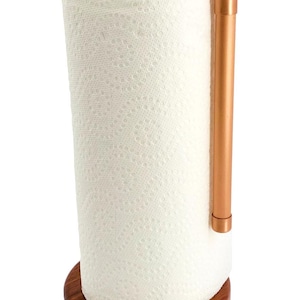 May include: A paper towel holder featuring a cylindrical dark brown wooden base and a vertical copper pipe arm that curves over the top to hold a roll of white paper towels in place. The design consists of a circular wood stand and metal pipe.
