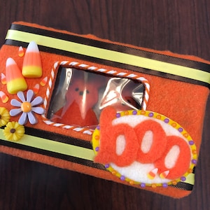 May include: Orange felt Halloween-themed box with a clear window, decorated with candy corn, flowers, and a "boo" applique. The box is trimmed with black and yellow ribbon, creating a festive and decorative storage item.