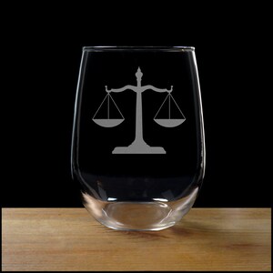 May include: A clear glass wine glass with a scale of justice etched on the side. The glass is sitting on a wooden surface.