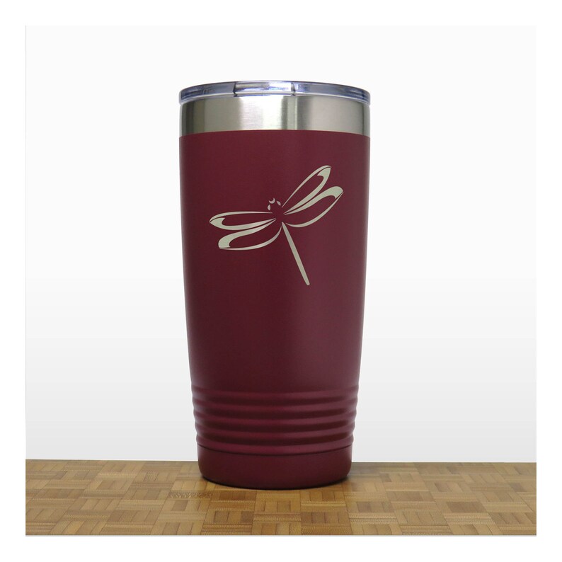 May include: A maroon stainless steel tumbler with a dragonfly design engraved on the side. The tumbler has a silver lid and a textured bottom.