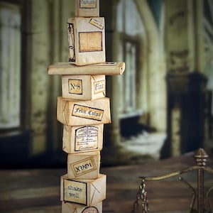 Puede incluir: Una pila de pequeñas cajas de aspecto envejecido con etiquetas, algunas con texto como "Optum", "Elixir" y "Caution!". Las cajas están dispuestas en una torre, lo que sugiere un tema de boticario o poción vintage. Una pequeña balanza es visible en el fondo.