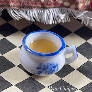 May include: A miniature white teacup with a blue rim and floral design, filled with a golden liquid. The tiny cup has a small handle and sits on a black and white checkered surface. The text "Petite Uniques" is visible.