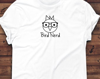 May include: White t-shirt with a black graphic of a bird wearing glasses and a crown. The text "Bird Nerd" is below the graphic. The shirt is displayed on a wooden surface.