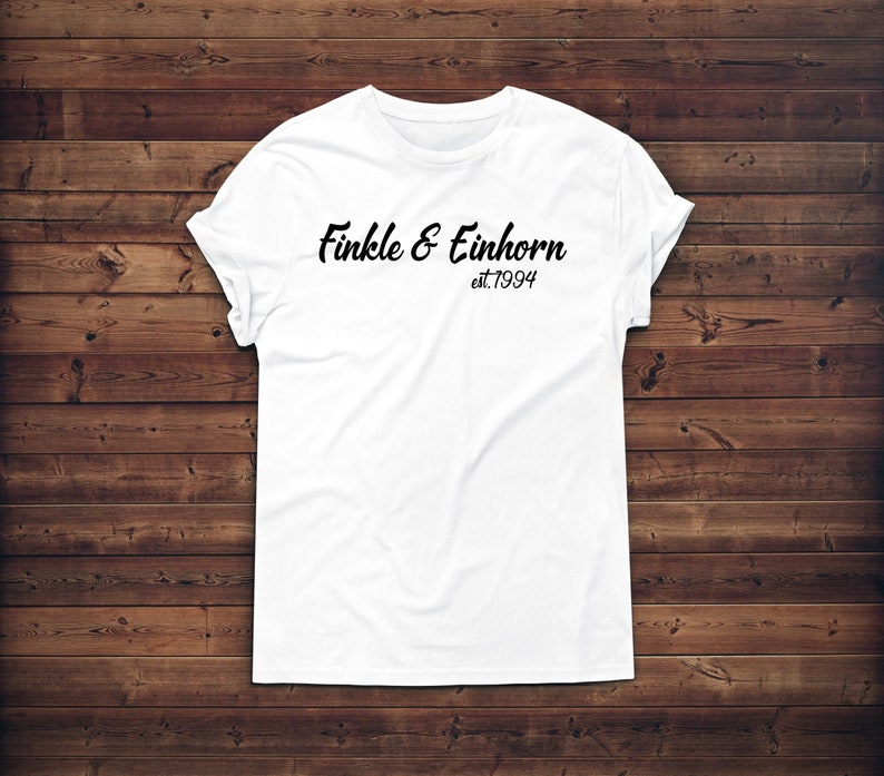 May include: A white t-shirt with the text "Finkle & Einhorn est.1994" in black script. The shirt is displayed on a wooden background. The sleeves are cuffed.
