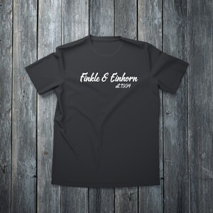 May include: A black t-shirt with the white text "Finkle & Einhorn est.1994" in a cursive font. The shirt is displayed on a dark wooden surface, creating a contrast with the light text.