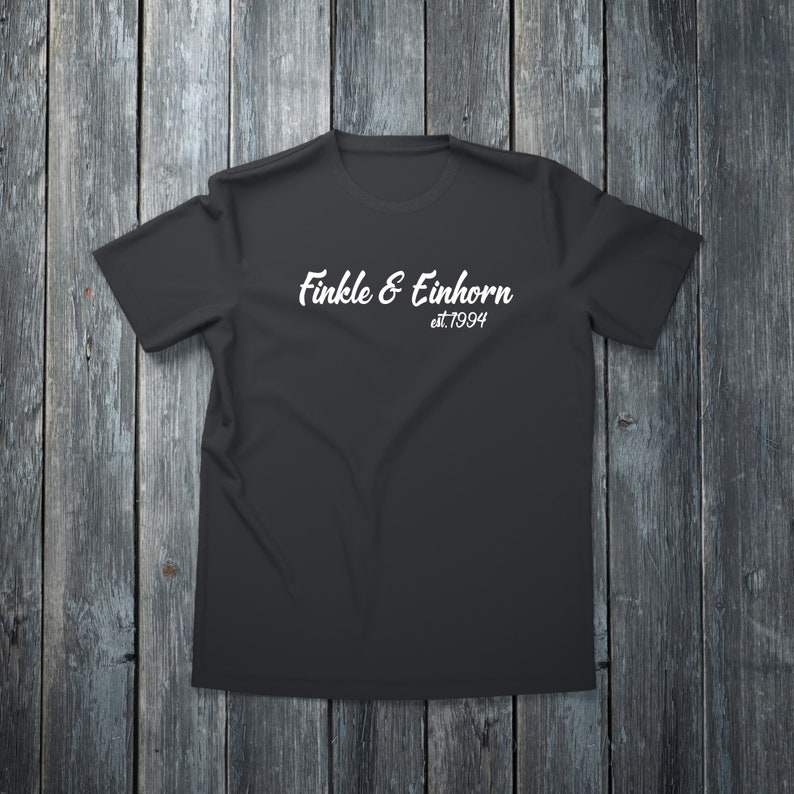 May include: A black t-shirt with the white text "Finkle & Einhorn est.1994" in a cursive font. The shirt is displayed on a dark wooden surface, creating a contrast with the light text.