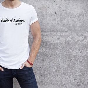 May include: White t-shirt with the text "Finkle & Einhorn at 1994" in black script. The shirt is worn by a person with their hands in their pockets, leaning against a gray concrete wall.