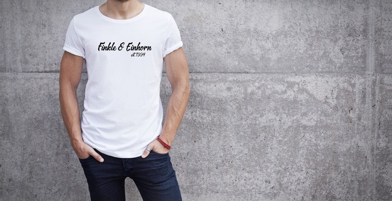 May include: White t-shirt with the text "Finkle & Einhorn at 1994" in black script. The shirt is worn by a person with their hands in their pockets, leaning against a gray concrete wall.