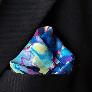 May include: A pocket square featuring a vibrant, swirling marble pattern in shades of bright blue, magenta, yellow, and deep purple. The fabric is folded into a triangular shape and tucked into the breast pocket of a solid black suit jacket.