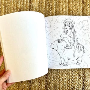 以下が含まれることがあります： 白紙のページと、カバに乗るキャラクターの白黒イラストが描かれた、開いた塗り絵。イラストは葉の模様で囲まれています。本は手で開かれています。